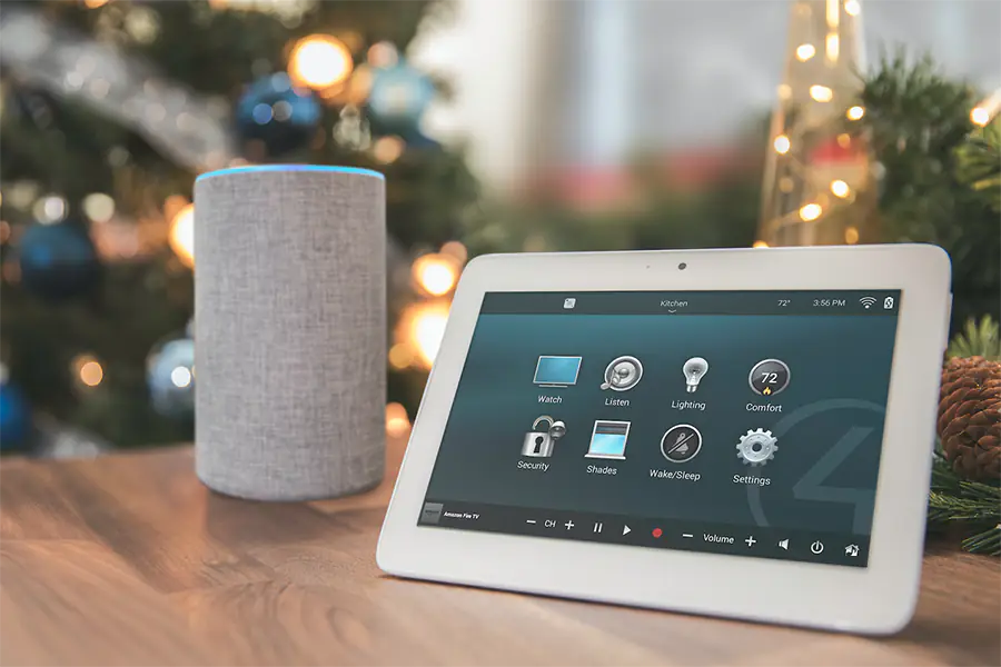 Tablet on table with Alexa speaker behind it. The tablet shows a menu with multiple settings, including Watch, listen, lighting, and comfort